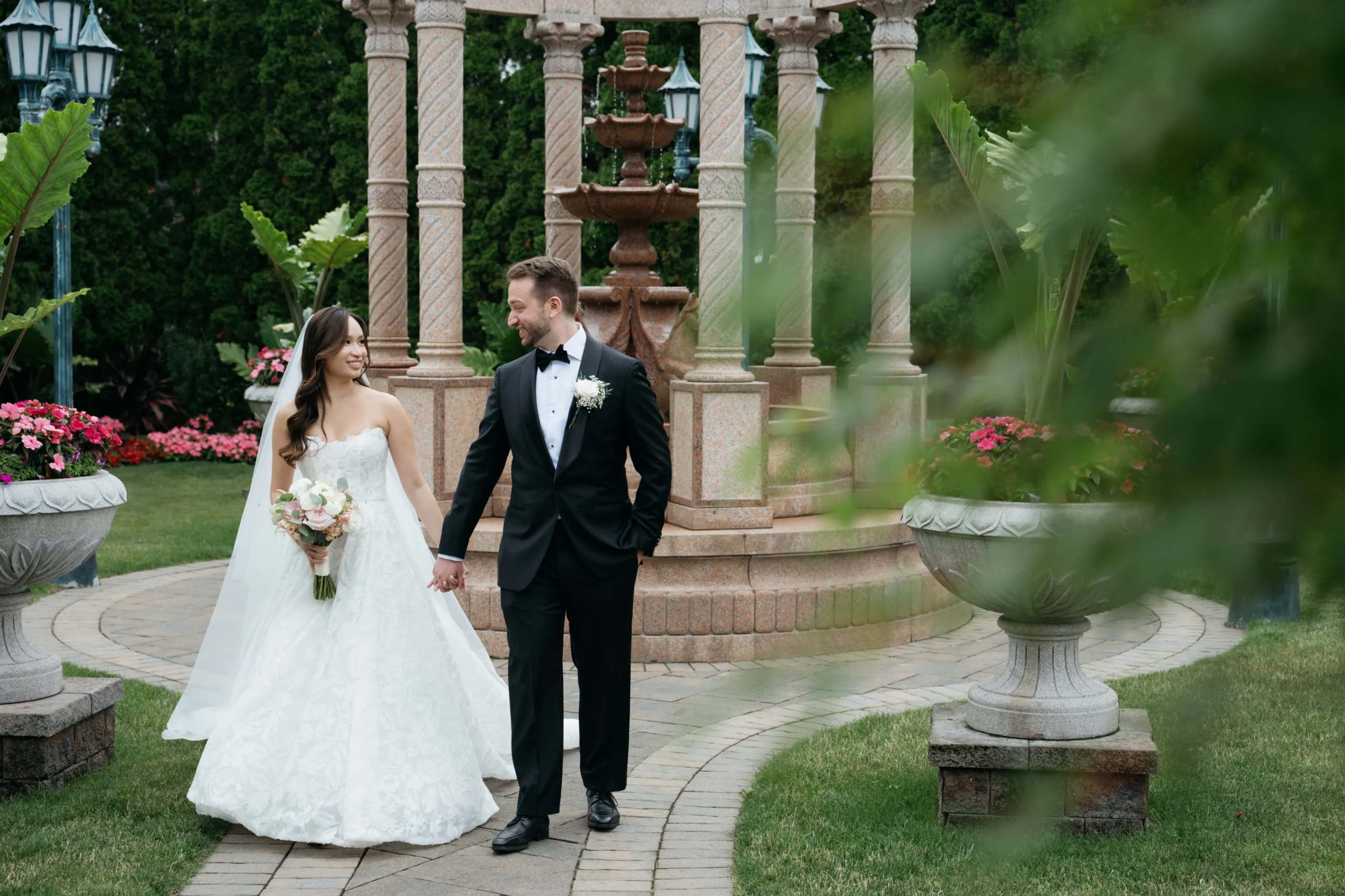 Newlywed couple walking hand in hand through a lush garden beside a stone fountain.