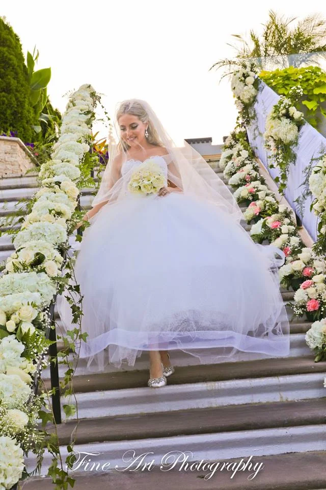 Bride walks down garden steps in a white wedding gown surrounded by flowers and greenery.