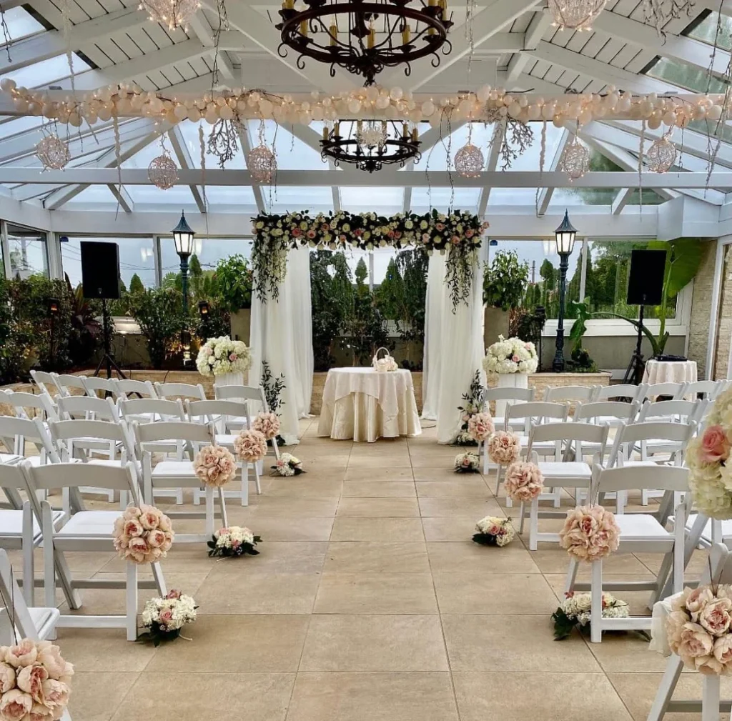 Indoor wedding ceremony in a glass atrium with rows of chairs, floral aisle arrangements, and a greenery covered altar.