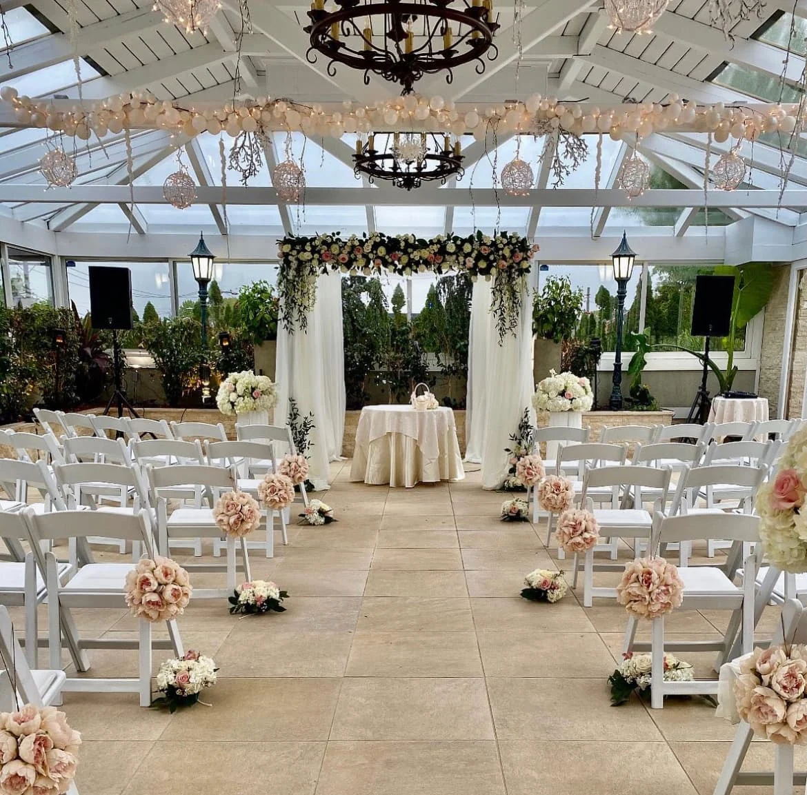 Indoor wedding ceremony in a glass atrium with rows of chairs, floral aisle arrangements, and a greenery covered altar.