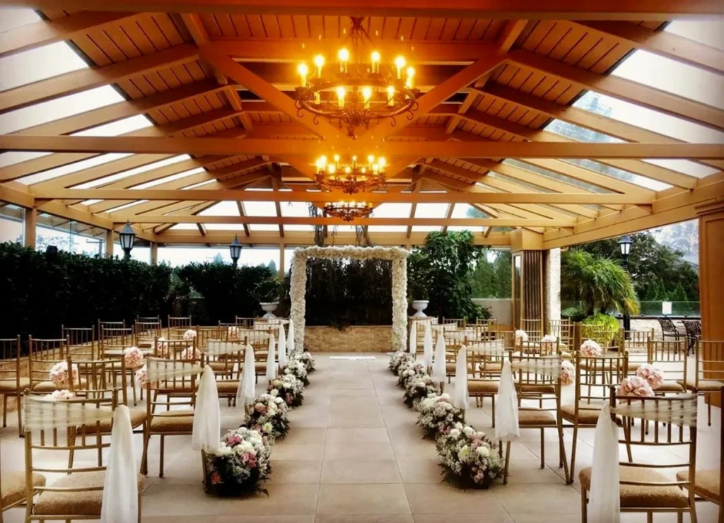 Indoor wedding ceremony setup with a flower lined aisle, wooden chairs, greenery, and a glowing chandelier under a wood beam ceiling.
