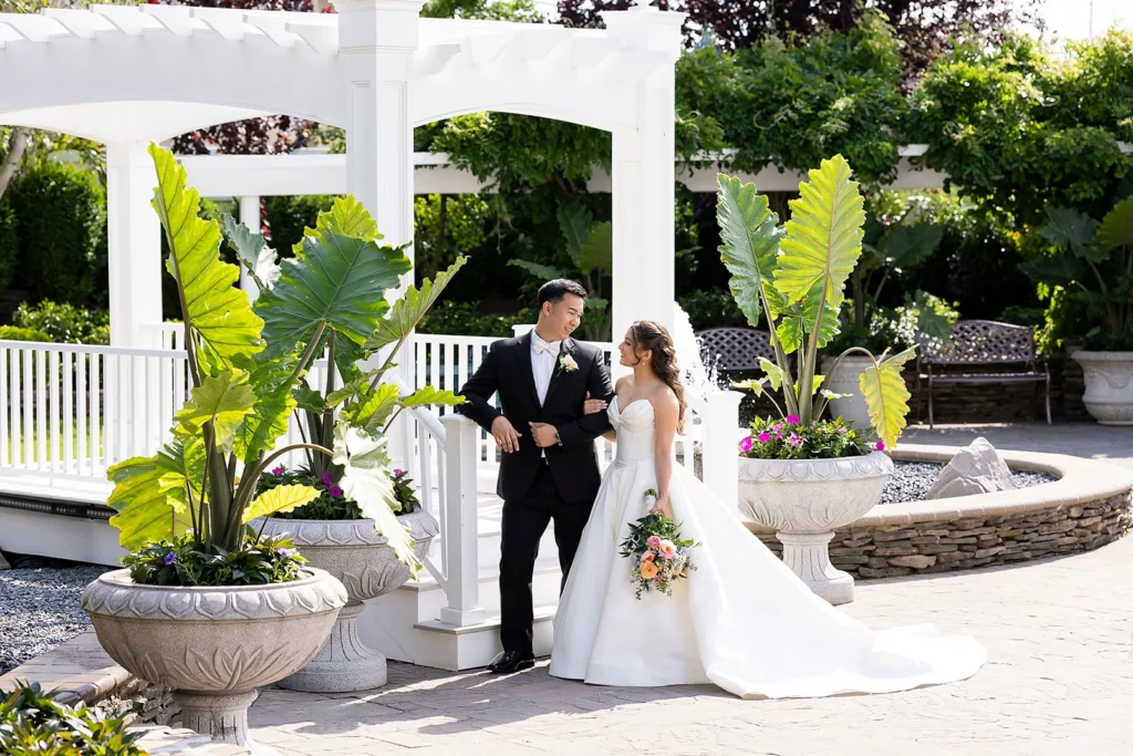 Bride and groom walk beside a white gazebo and fountain in a lush outdoor wedding garden.