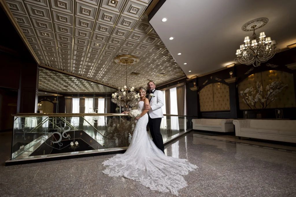 Bride and groom pose in an elegant ballroom foyer with chandeliers, patterned carpeting, and ornate ceiling details.