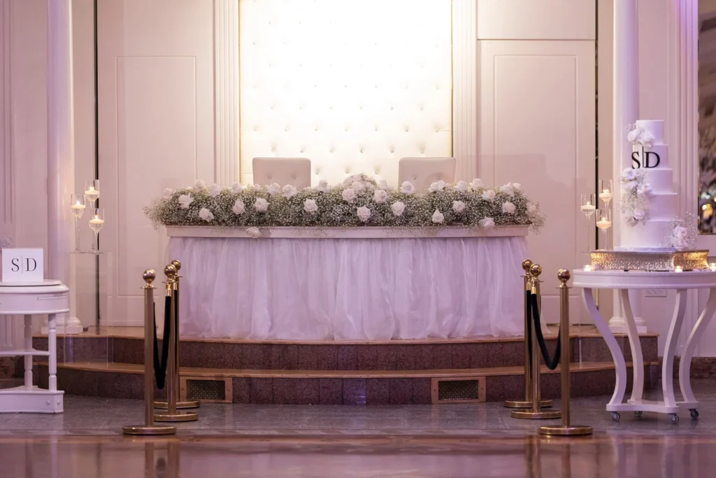 Elegant wedding sweetheart table with floral arrangements, white draping, candles, and soft pink uplighting.