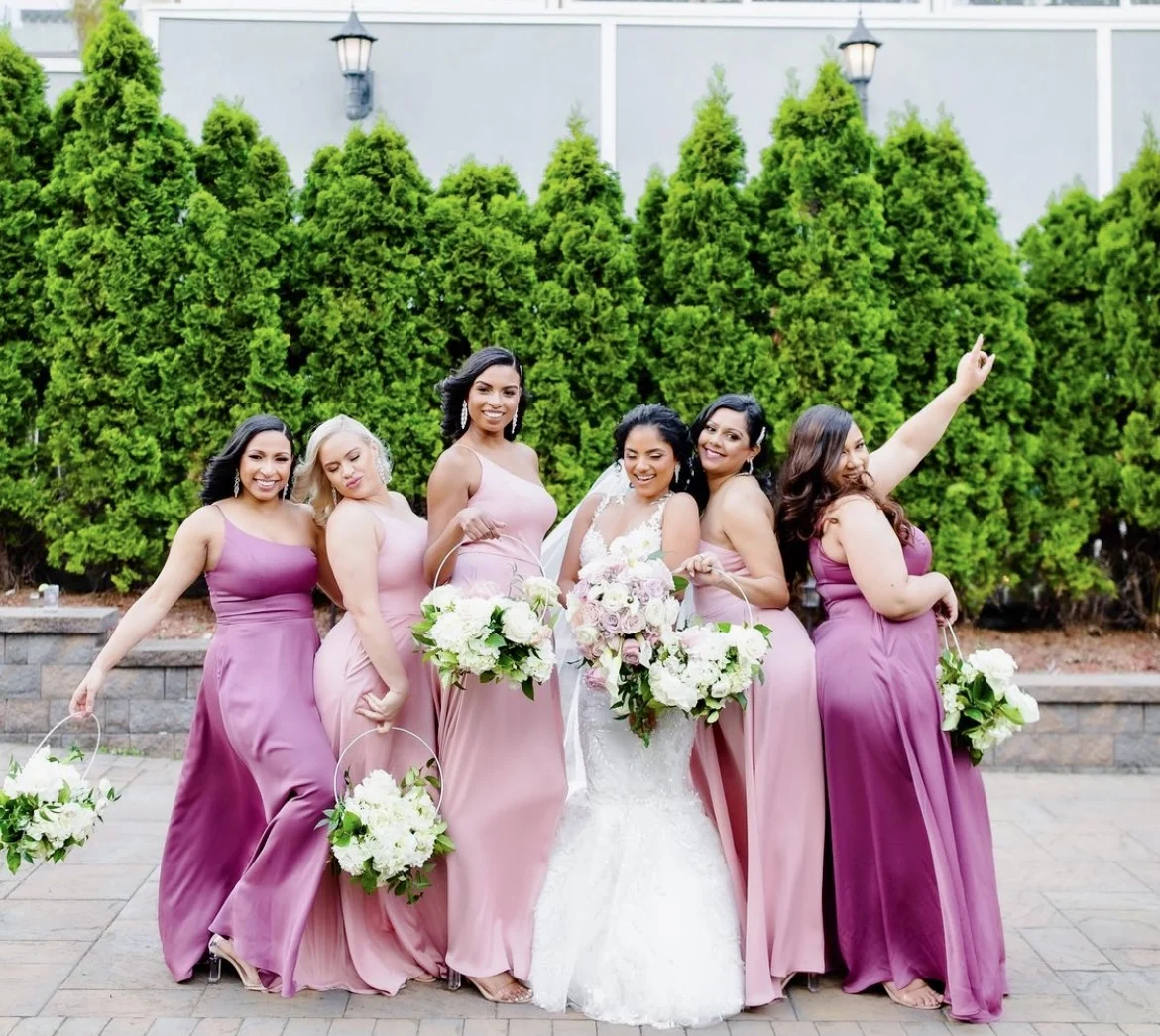 Bride in a white wedding gown celebrating outdoors with bridesmaids in pink dresses and bouquets.